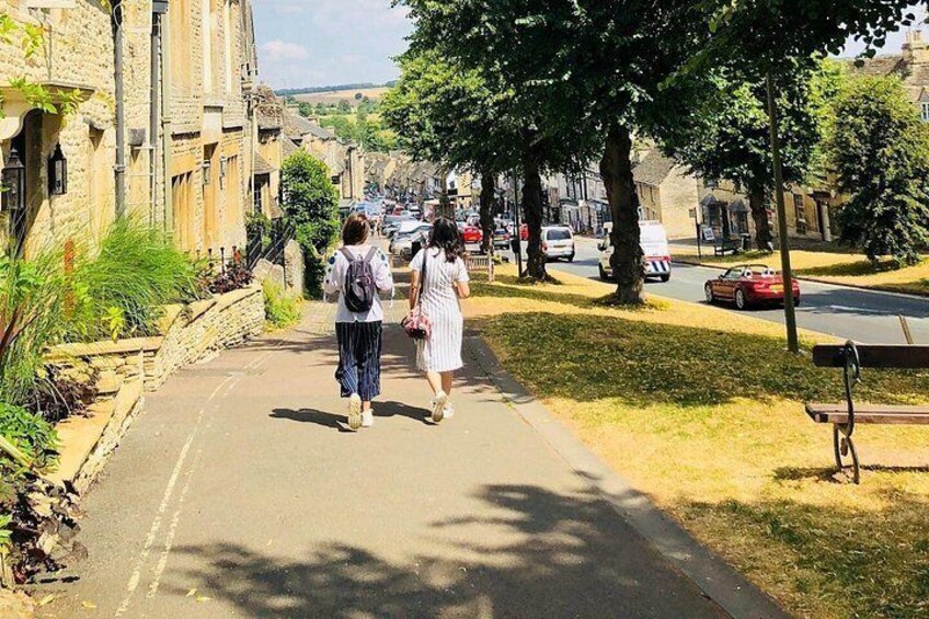 A paved pathway with two people walking, surrounded by stone buildings and trees. 1