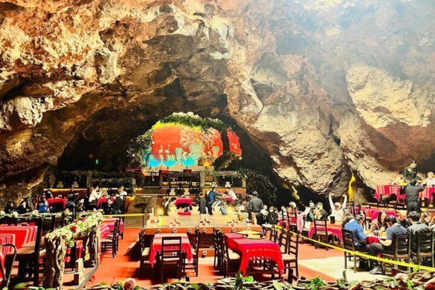 An indoor dining area with tables and chairs, a stage with a colorful backdrop, and a cave-like rock formation in the background. 1