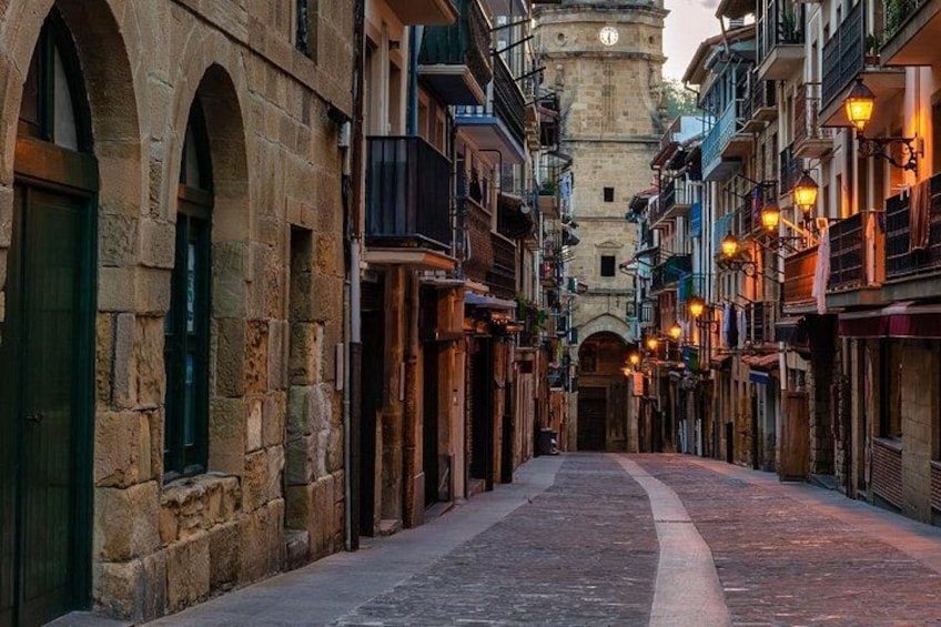 A narrow street with stone buildings, a clock tower, and warm lighting. 2