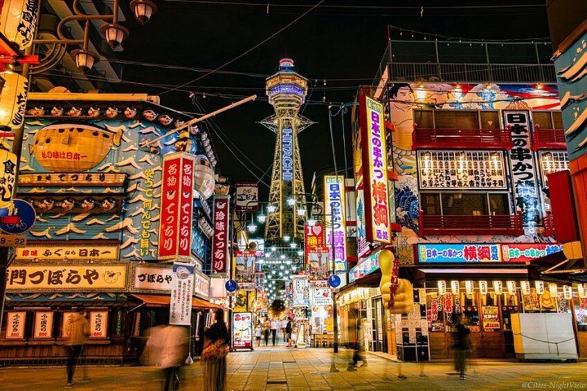 A brightly lit street with traditional Japanese signage and a prominent tower in the background. 1