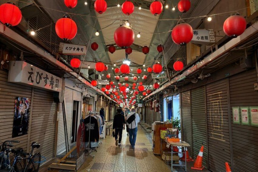 A narrow alley with red lanterns hanging from the ceiling, closed shops, and a few people walking. 4