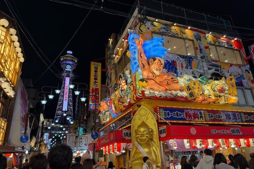 A brightly lit street with a large illuminated building featuring traditional Japanese artwork and characters. 3