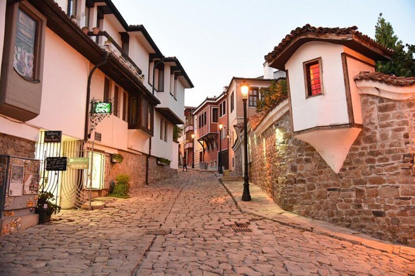 Una calle empedrada bordeada por edificios tradicionales, un muro de piedra y un farol. 4