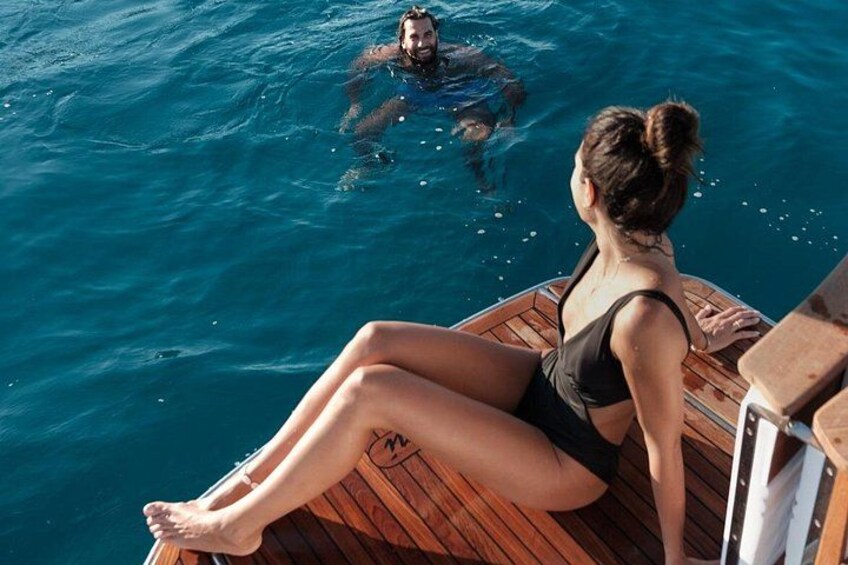 Une femme assise sur le pont en bois d’un bateau, un homme qui nage dans l’eau et une mer d’un bleu limpide. 2