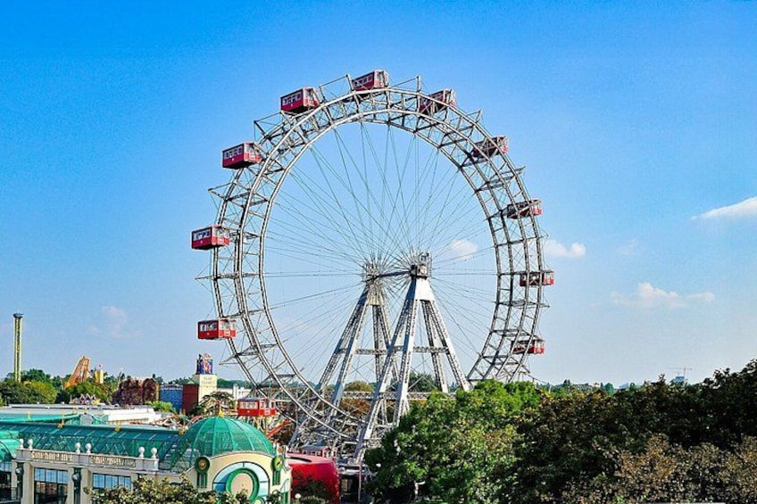 A large Ferris wheel with cabins, a green building with a dome, and trees in the foreground. 4