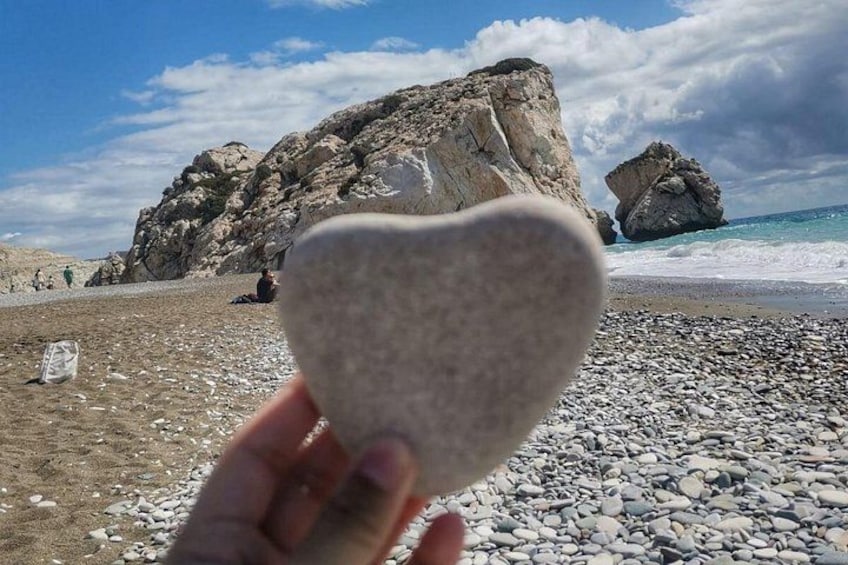 A hand holding a heart-shaped stone on a pebbly beach with large rocks and the ocean in the background. 2