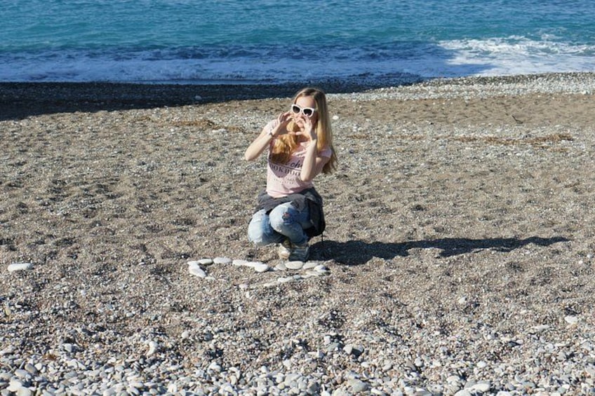 A person crouching on a pebbled beach with the ocean in the background. 5