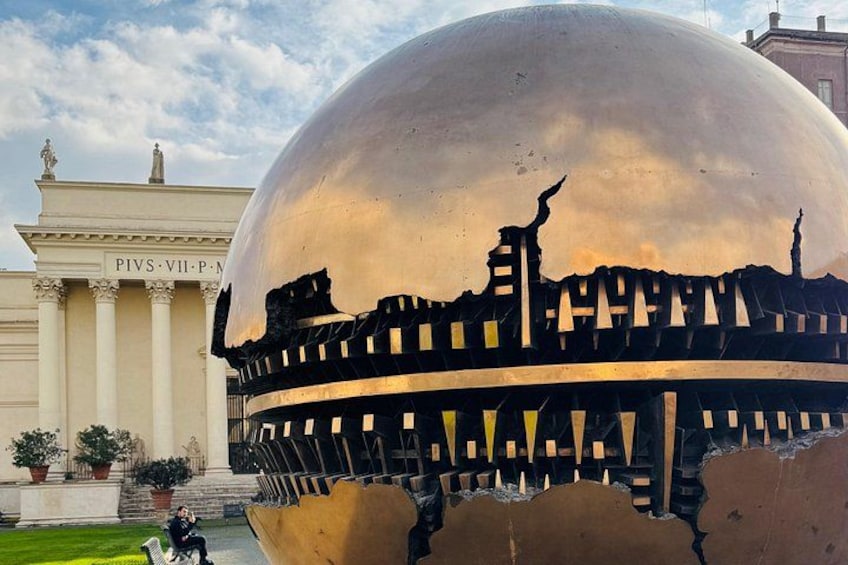 A large metallic sphere with a cracked surface, reflecting a cityscape. In the background, there's a building with columns and a sign that reads 'PIVS-VII-P. M.' 5