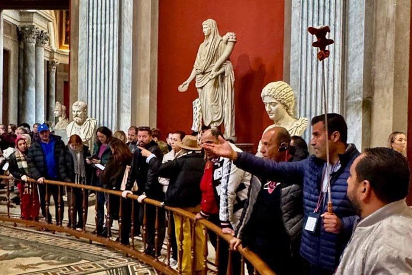 Un grupo de personas hacen fila, algunas sosteniendo pancartas, en una sala con arquitectura clásica y estatuas. 3