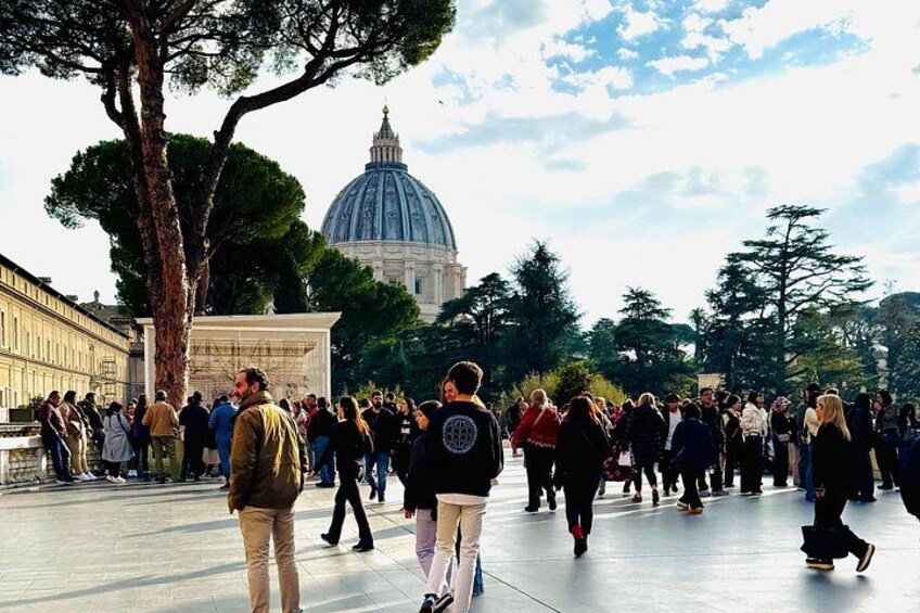 A crowded square with people walking, a large tree, and a dome-shaped building in the background. 2