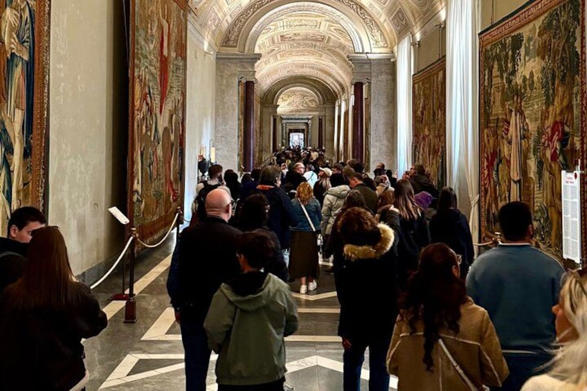 A crowded hallway with people walking, large paintings on the walls, and a marble floor. 3