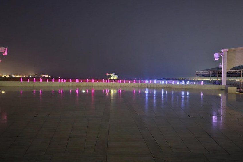 A nighttime scene with a waterfront promenade, illuminated fountains, and a modern building structure. 5