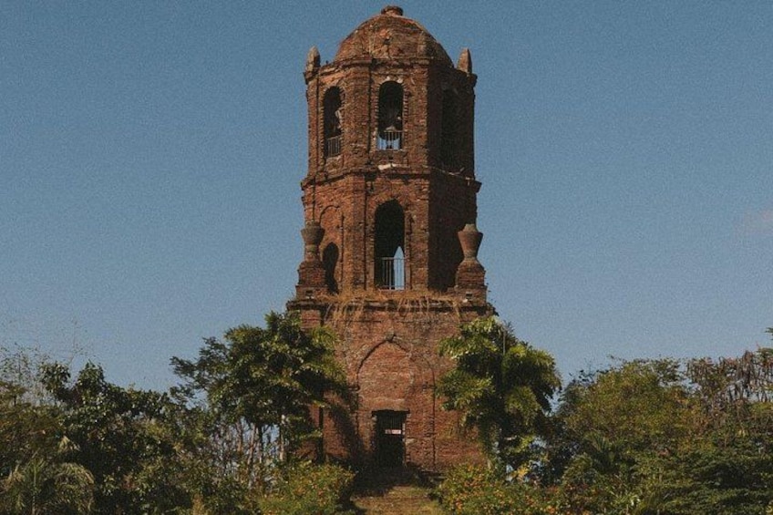 Ein hoher, roter Backturm mit Bogenfenstern und einer Kuppel, umgeben von üppigem Grün. 3