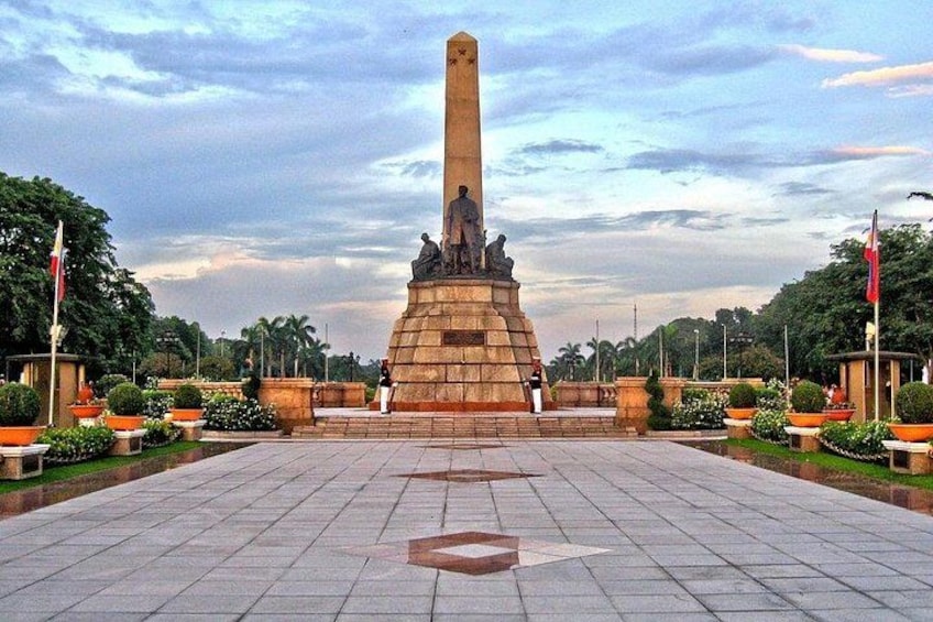 Ein großes Denkmal mit einer Statue, umgeben von einem gepflasterten Platz mit Blumenbeeten und Bäumen. 4