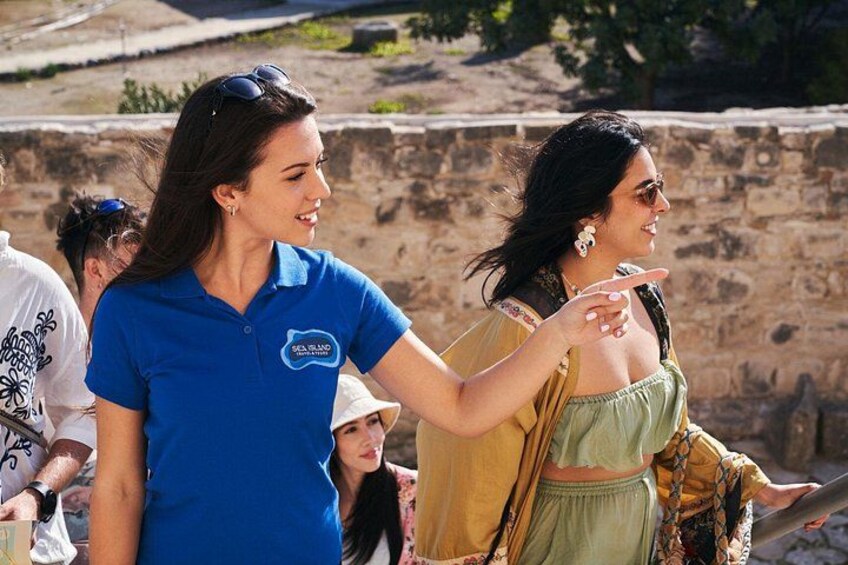 A group of people outdoors, some wearing casual clothing, with a stone wall and greenery in the background. 3