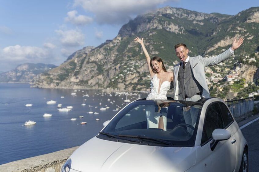 A couple in a convertible car with a coastal landscape and mountains in the background. 4