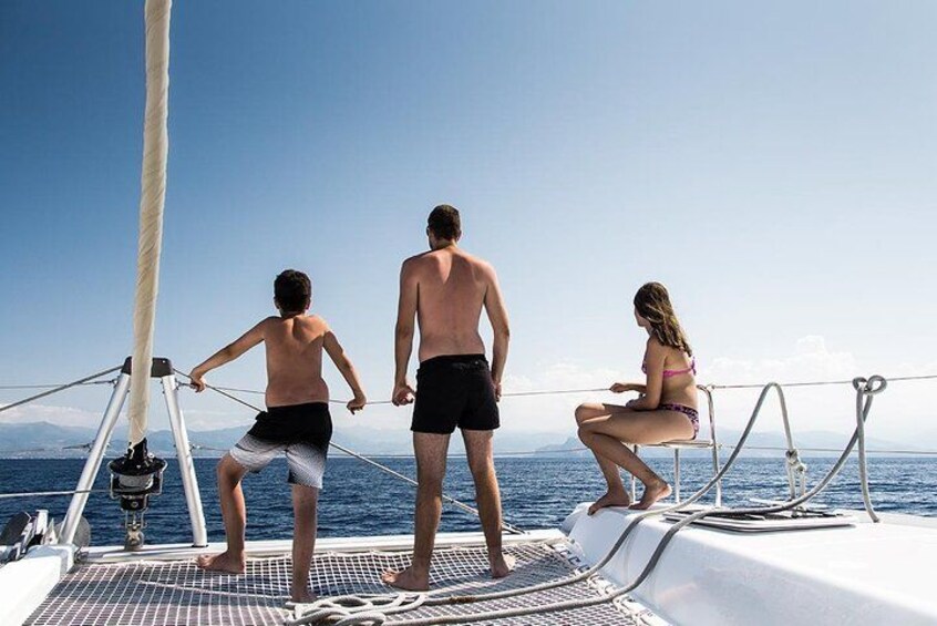Three people on a sailboat, one standing and two sitting, with a clear sky and ocean in the background. 5