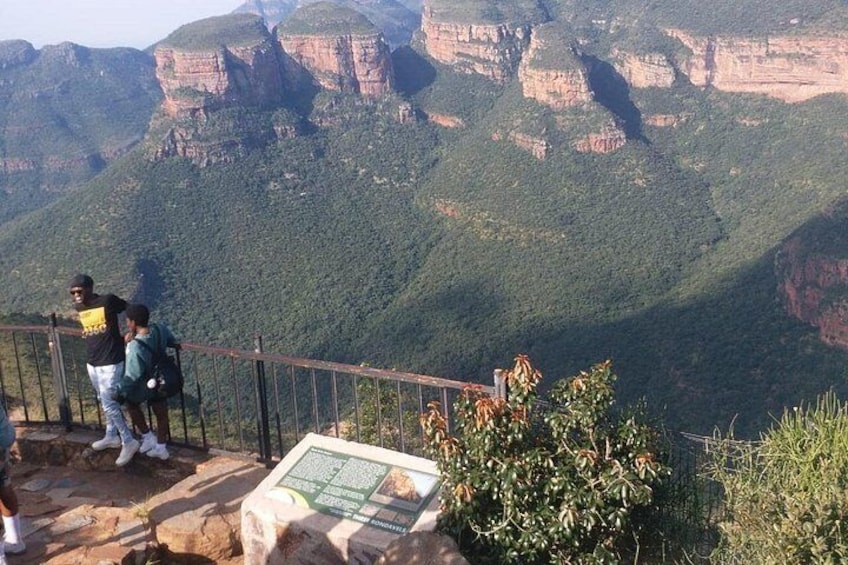 A mountainous landscape with a viewing platform, people observing the scenery, and a sign with information. 1