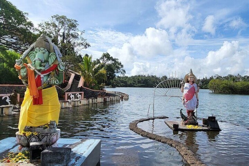 Une statue d’une divinité, placée sur un piédestal au milieu d’une étendue d’eau, entourée de verdure et sous un ciel dégagé. 1