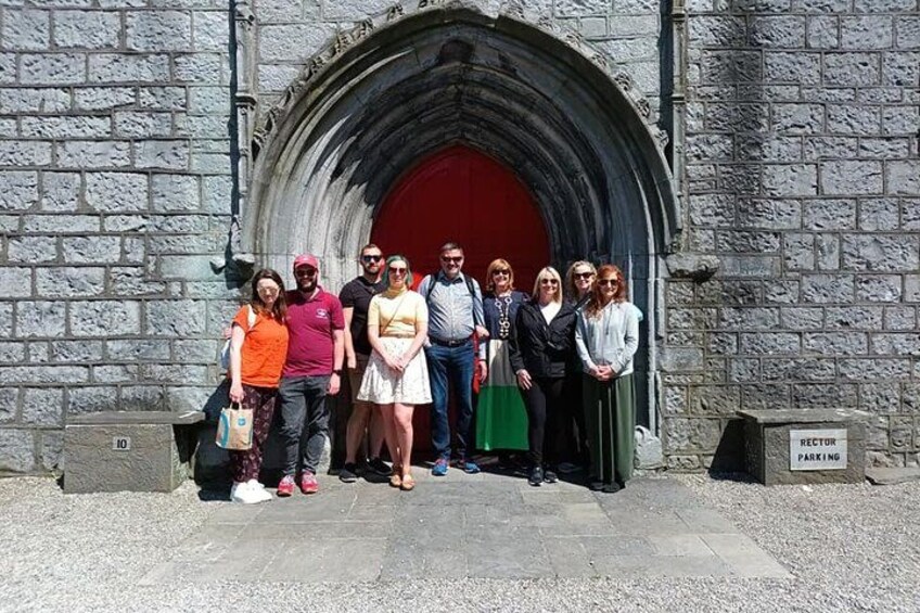 Un groupe de personnes se tient devant un bâtiment en pierre, qui possède une grande porte d’entrée en arc de cercle. 1