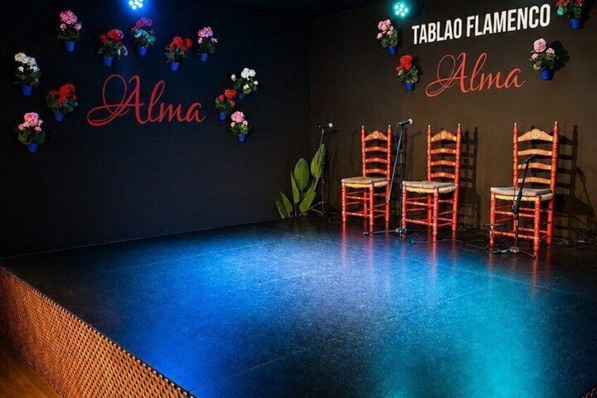 A stage with chairs, a wall with flowers, and the text 'TABLAO FLAMENCO Alma'. 5