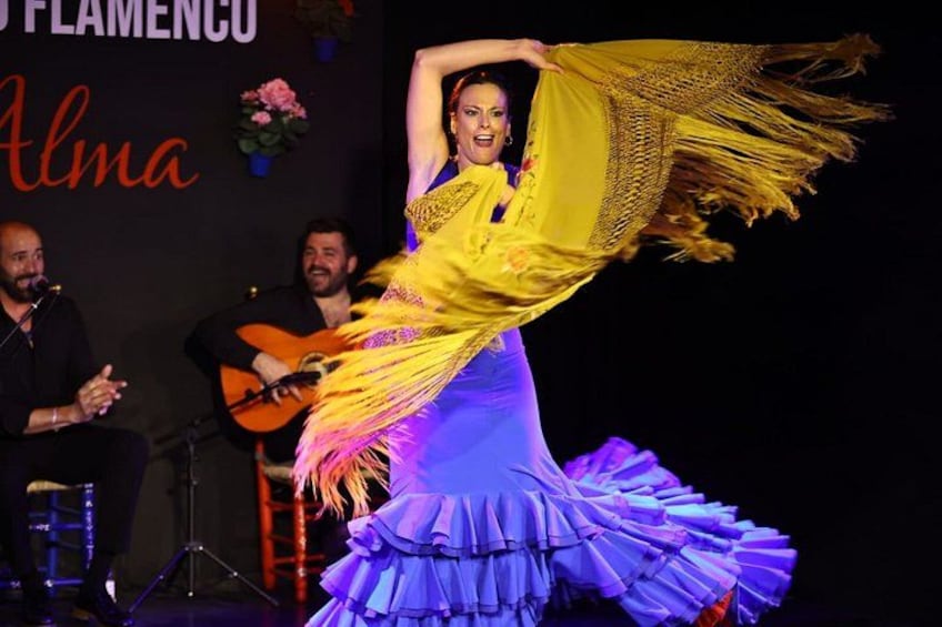 A flamenco dancer in a vibrant yellow and purple dress performing on stage with a guitar player in the background. 1