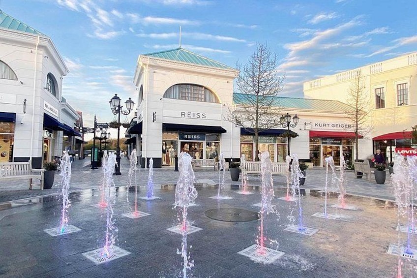 A shopping plaza with fountains, Reiss and Kurt Geiger stores, and a clear sky. 4