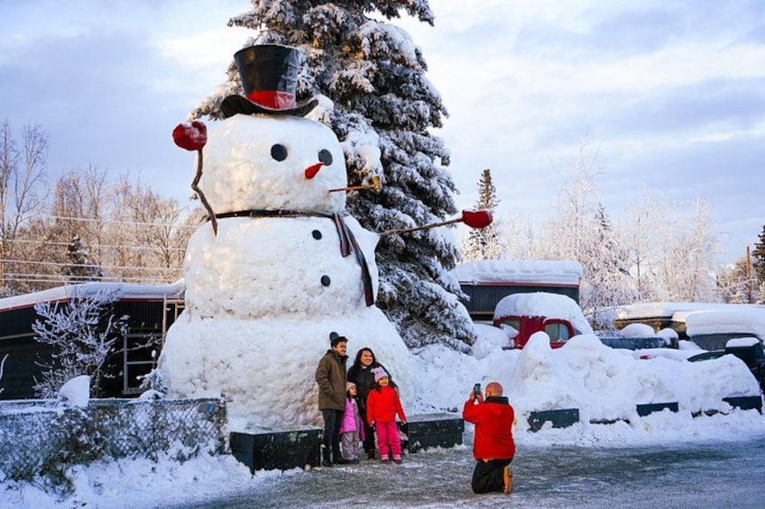 Ein großer Schneemann mit Zylinder und Karottennase, ein mit Schnee bedeckter Baum und geparkte Autos auf einem verschneiten Parkplatz. 1