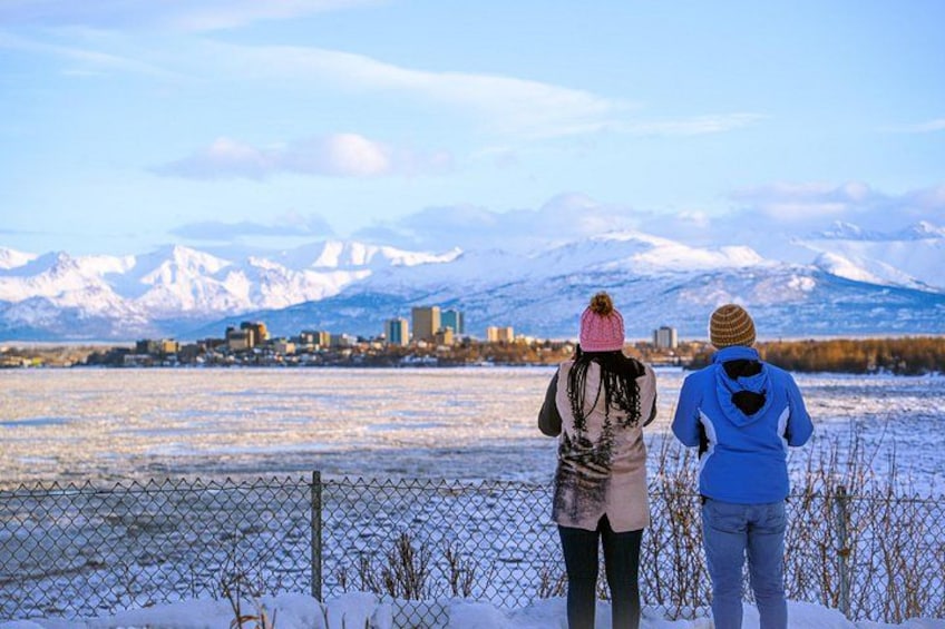 Zwei Personen stehen an einem Zaun und blicken auf eine verschneite Landschaft mit Bergen im Hintergrund. 4