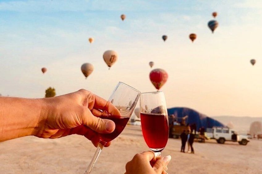 Due mani che sorreggono calici di vino, con un paesaggio desertico e mongolfiere sullo sfondo. 2