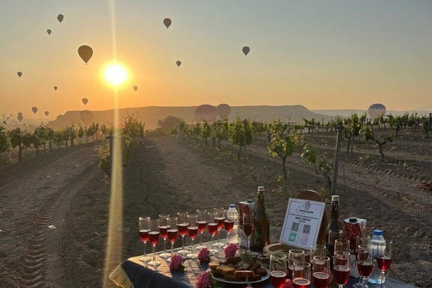 Un vigneto con un tavolo apparecchiato per un pasto, bottiglie di vino e bicchieri. Si vedono palloni aerostatici nel cielo. 3