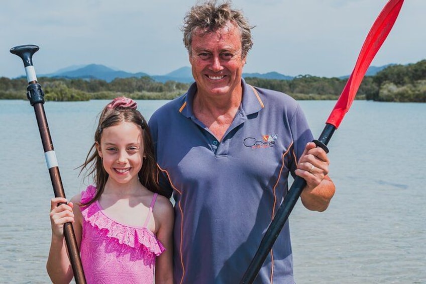A man and a young girl holding paddles on a calm lake with mountains in the background. 3