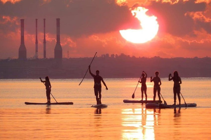 Personas practicando paddleboard en un cuerpo de agua al atardecer, con estructuras industriales al fondo. 4