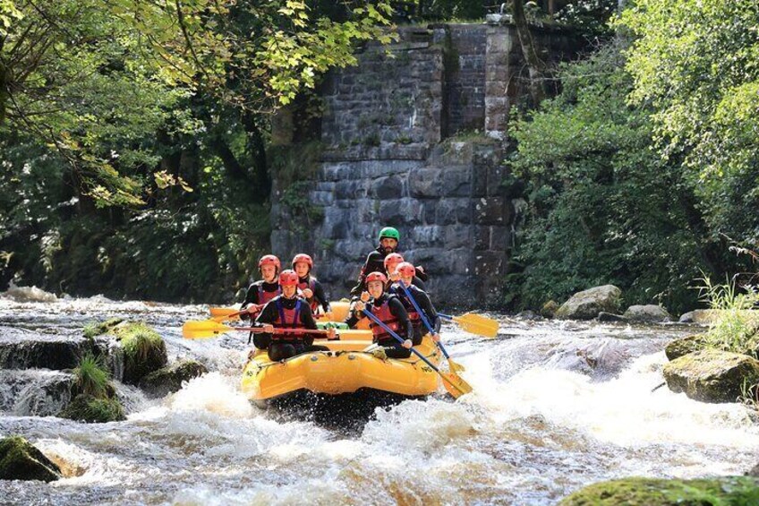 A group of people rafting on a yellow raft down a river with a stone bridge in the background. 2