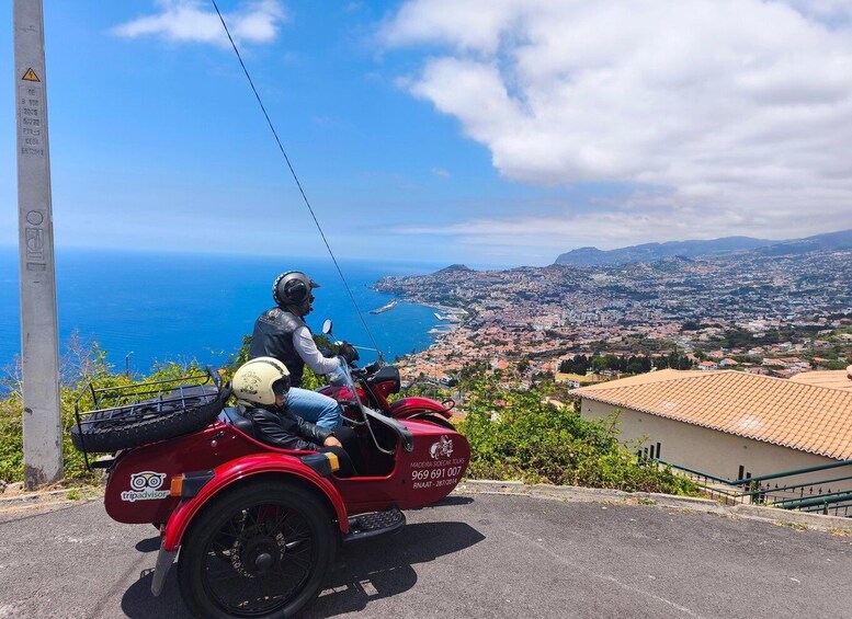 Una motocicleta roja con sidecar estacionada en una carretera, con vistas a un paisaje urbano costero bajo un cielo azul despejado. 1