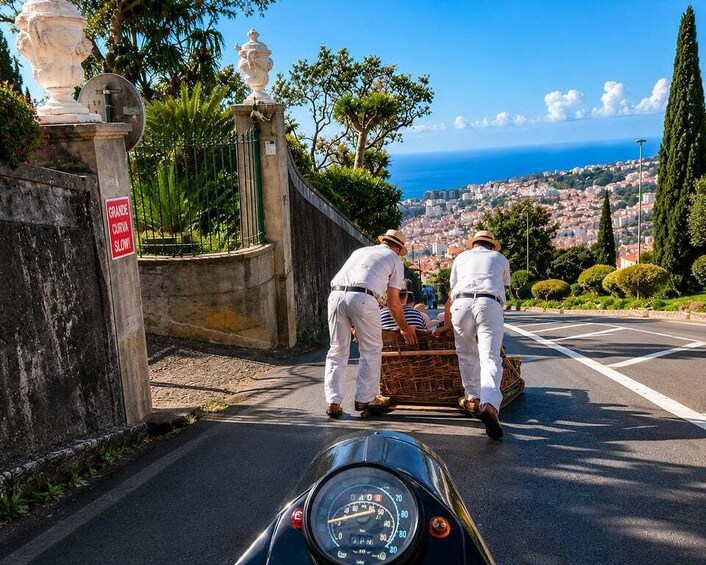 Dos hombres con uniformes blancos empujan un carrito por un camino, con vistas a un pueblo costero al fondo. 2