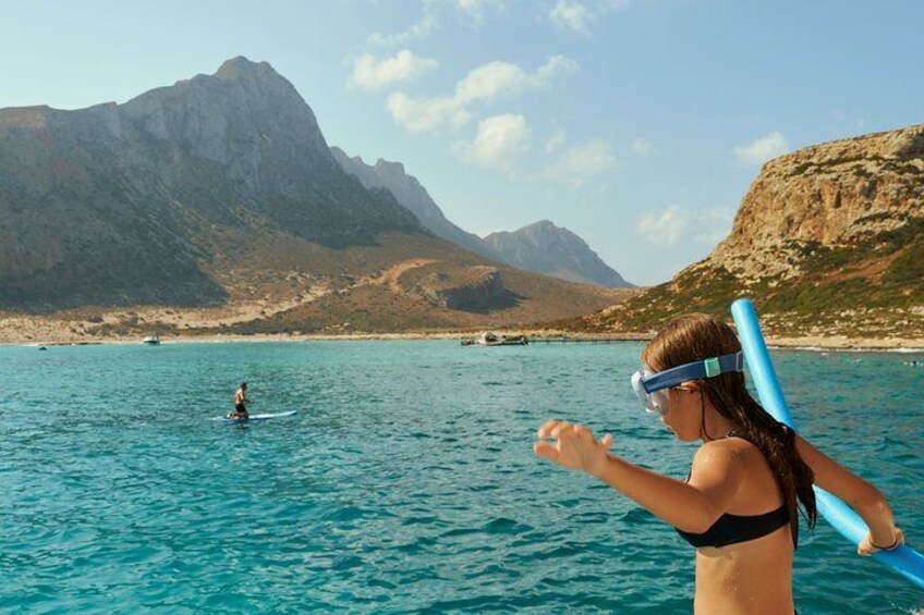 A person in a black swimsuit holding a blue snorkel, standing in a turquoise sea with a mountainous landscape in the background. 5