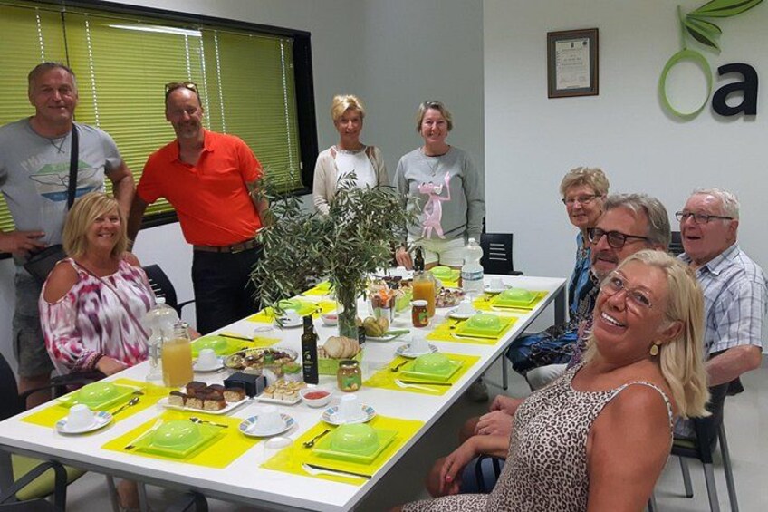 A group of people seated around a table with food and drinks, in a room with a plant and a wall decoration. 2