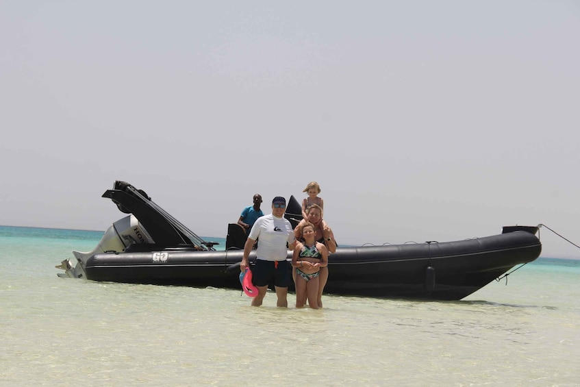 A black inflatable boat with people standing on the shore, a clear turquoise sea, and a clear sky. 4