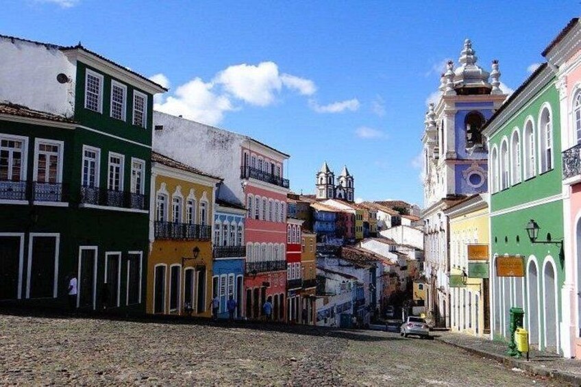A cobblestone street lined with colorful buildings, a church with a bell tower, and a clear blue sky. 4