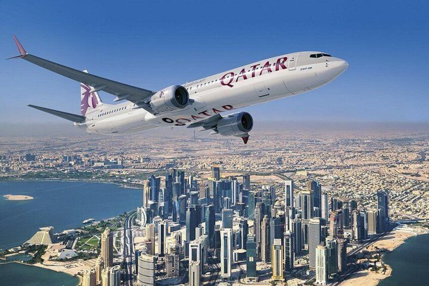 Ett Qatar Airways-flygplan flyger över en stad med höga byggnader och en kustlinje. 1