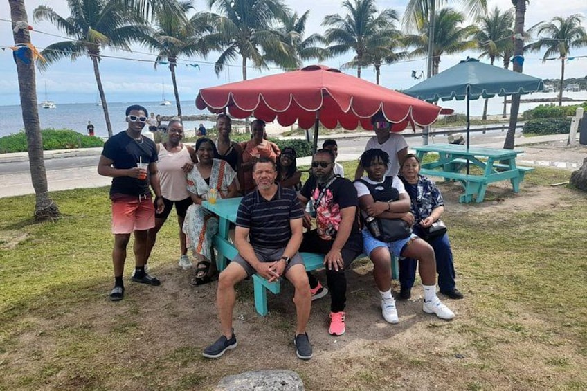 Un grupo de personas sentadas y de pie cerca de una playa, con palmeras y una sombrilla roja al fondo. 3