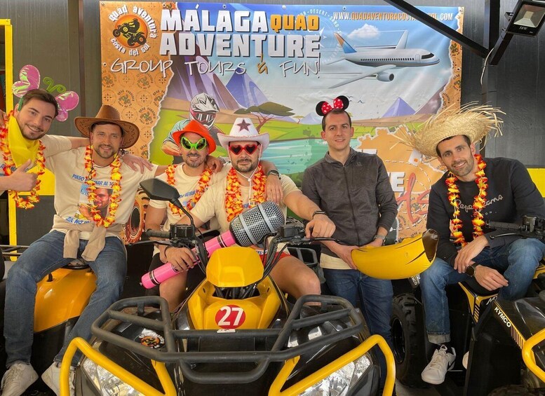 En grupp personer på en fyrhjuling, klädda i festliga blomsterhalsband, med en skylt med texten ”Malaga Quad Adventure” och en illustration av ett ökenlandskap i bakgrunden. 1