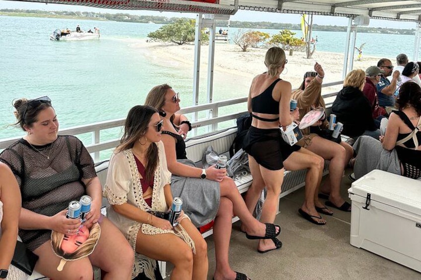 People seated on a boat, some holding drinks, with a beach and water in the background. 1