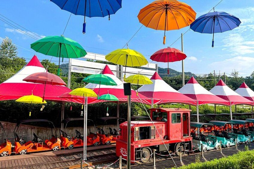 A train station with colorful umbrellas, a red train, and a row of parked cars. 2