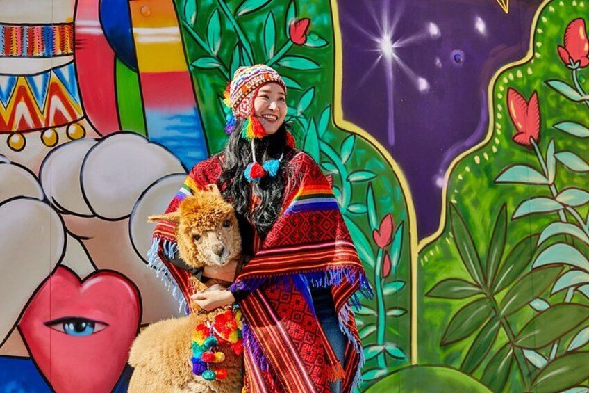 Una persona vestida con traje tradicional sosteniendo una llama, con un mural colorido de fondo. 2