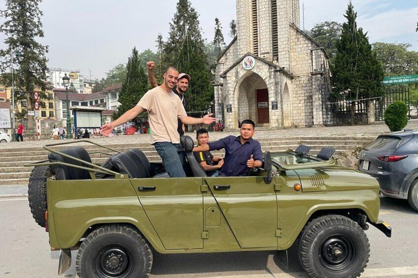 A green military-style vehicle with three people on it, parked in front of a church with a tall steeple. 3