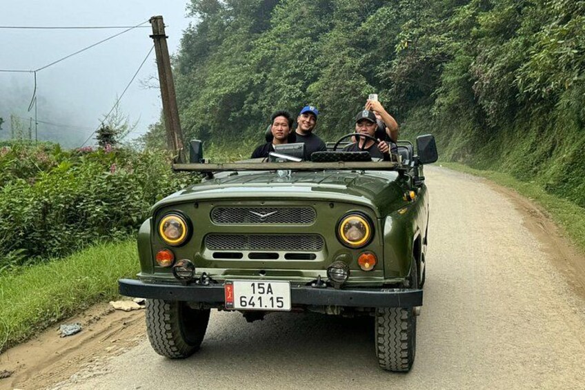 A green military vehicle on a rural road with two passengers, surrounded by greenery. 4