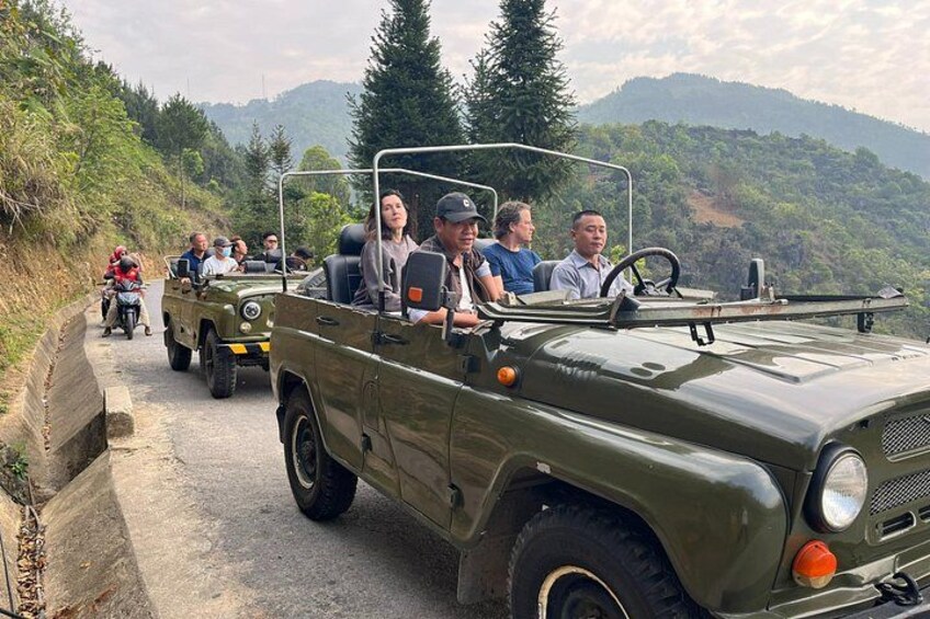A group of people in a green jeep on a narrow road surrounded by greenery and hills. 2