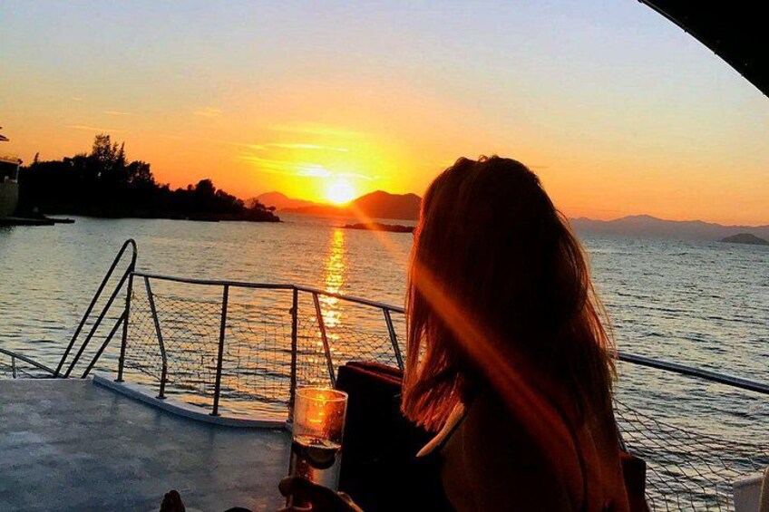 Solnedgang over vand med et gelænder, og en person holder et glas med drik. 1